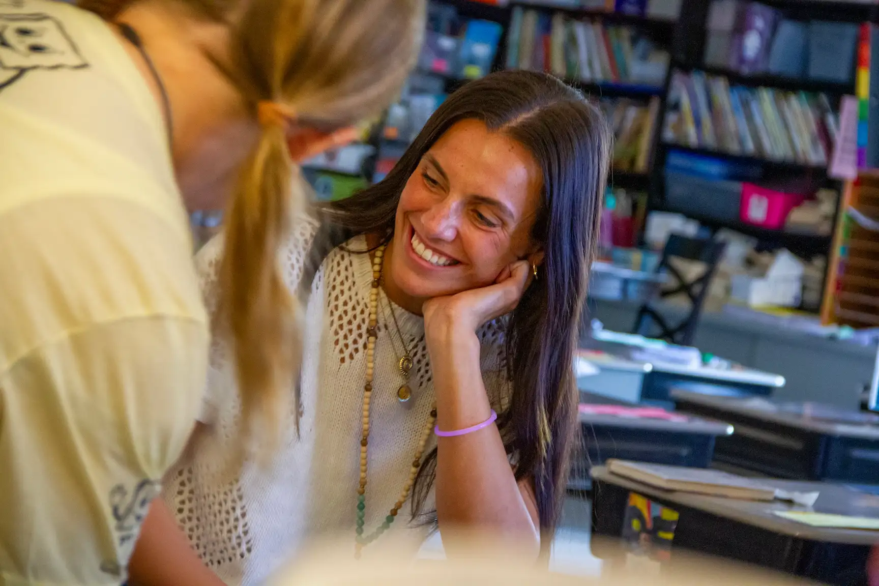 A woman works in a classroom, smiling at a student to the left. Bookshelves and desks are visible in the background.